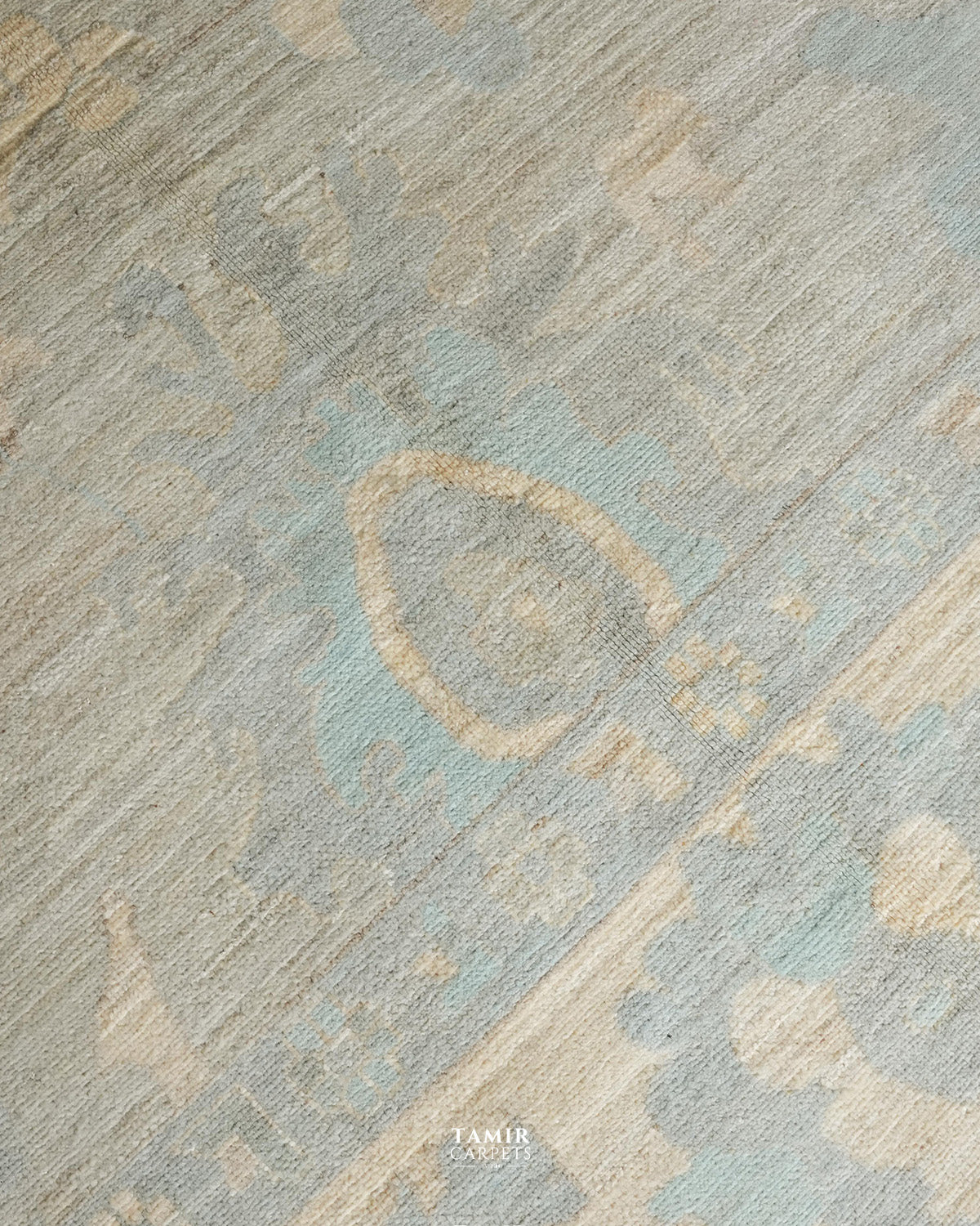 A close-up of a textured area rug featuring a distressed, abstract pattern in muted shades of light blue, beige, and gray. A small 'TAMIR CARPETS' brand watermark is visible at the bottom center.