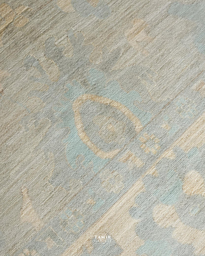 A close-up of a textured area rug featuring a distressed, abstract pattern in muted shades of light blue, beige, and gray. A small 'TAMIR CARPETS' brand watermark is visible at the bottom center.