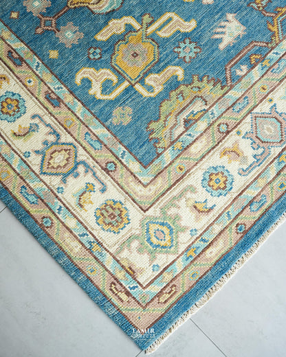 A close-up corner view of a traditional patterned area rug with a blue field, cream borders, and accents of yellow, beige, and brown, laid on a light gray floor.