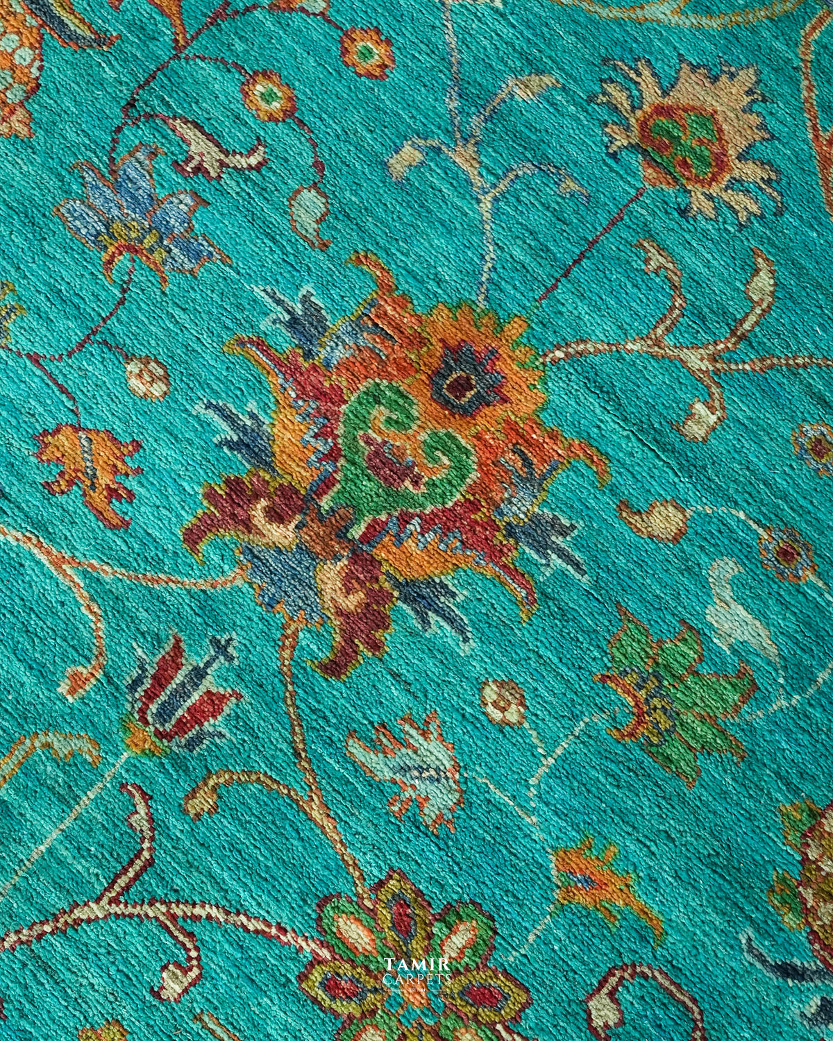 A close-up of a woven rug or textile with a turquoise blue background and an ornate floral pattern in orange, green, red, beige, and blue. The texture is highly visible, and a small 'TAMIR CARPETS' brand watermark is visible near the bottom center.
