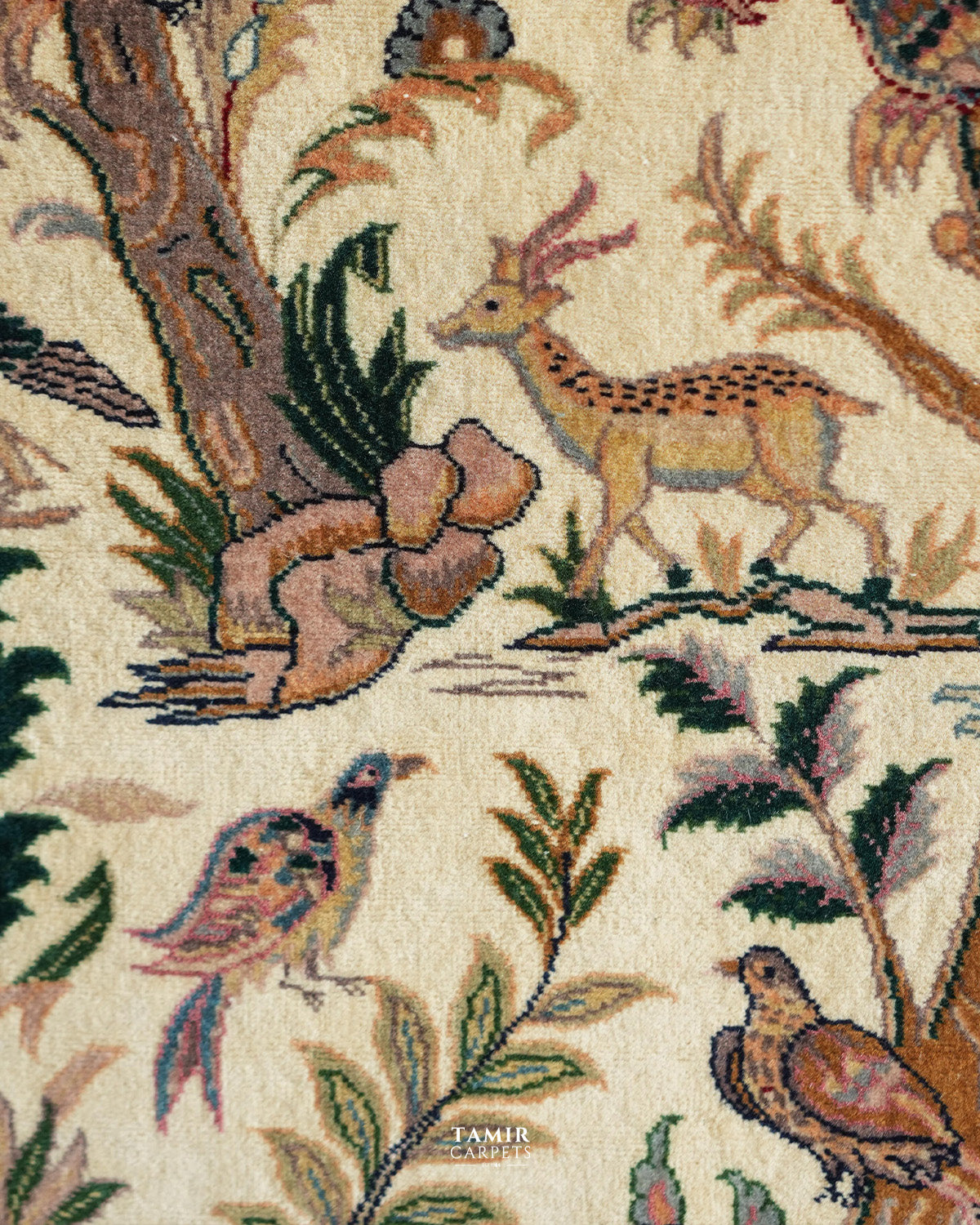 Close-up of a woven rug or textile with a naturalistic woodland pattern featuring a deer, birds, foliage, and tree trunk on a warm cream background. The brand name TAMIR CARPETS is visible near the bottom center.