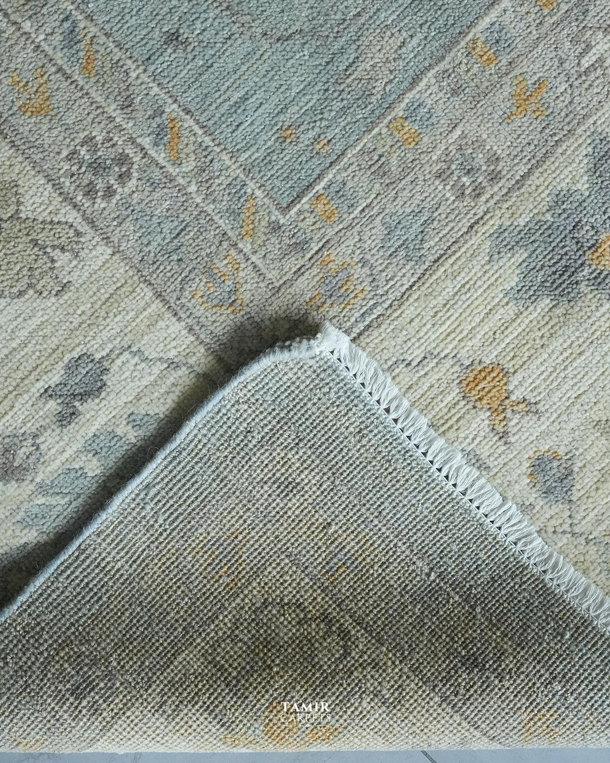 A close-up of a patterned area rug showing its corner and fringe detail. The rug features a distressed, vintage-inspired design in muted tones of slate blue, beige, gray, and ochre yellow.