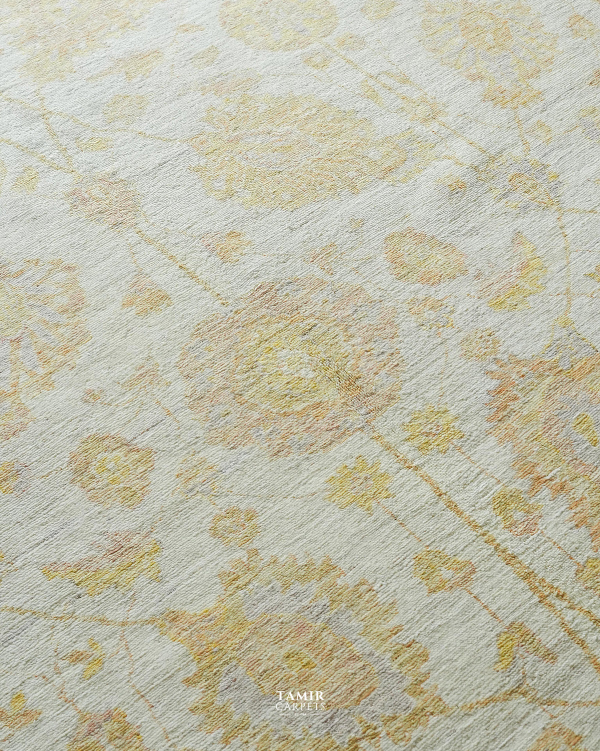 A close-up of a textured area rug featuring a delicate, faded floral pattern in soft yellow and muted peach tones over an off-white base. The brand name 'TAMIR CARPETS' is printed in white text at the bottom center.