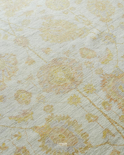 A close-up of a textured area rug featuring a delicate, faded floral pattern in soft yellow and muted peach tones over an off-white base. The brand name 'TAMIR CARPETS' is printed in white text at the bottom center.