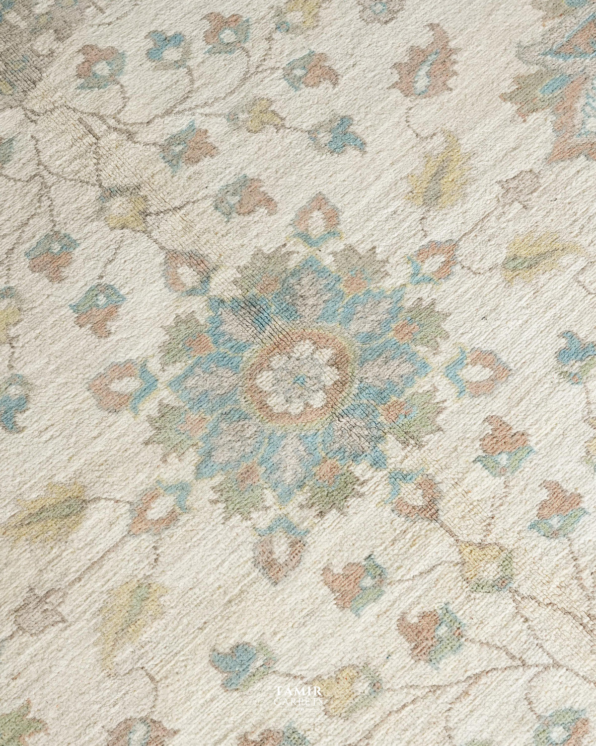 A close-up of a textured area rug featuring a traditional floral and medallion pattern in muted tones of blue, beige, and brown on a cream background.