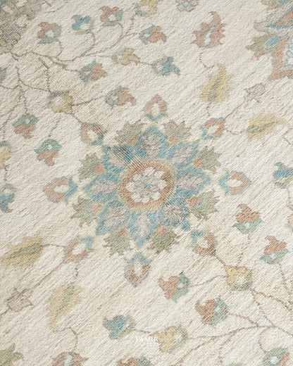 A close-up of a textured area rug featuring a traditional floral and medallion pattern in muted tones of blue, beige, and brown on a cream background.