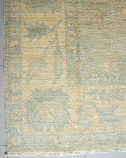 A close-up of a traditional handwoven rug with a light beige base and muted sage green and dusty pink patterns, featuring a fringed edge and a visible TAMIR CARPETS logo at the bottom.