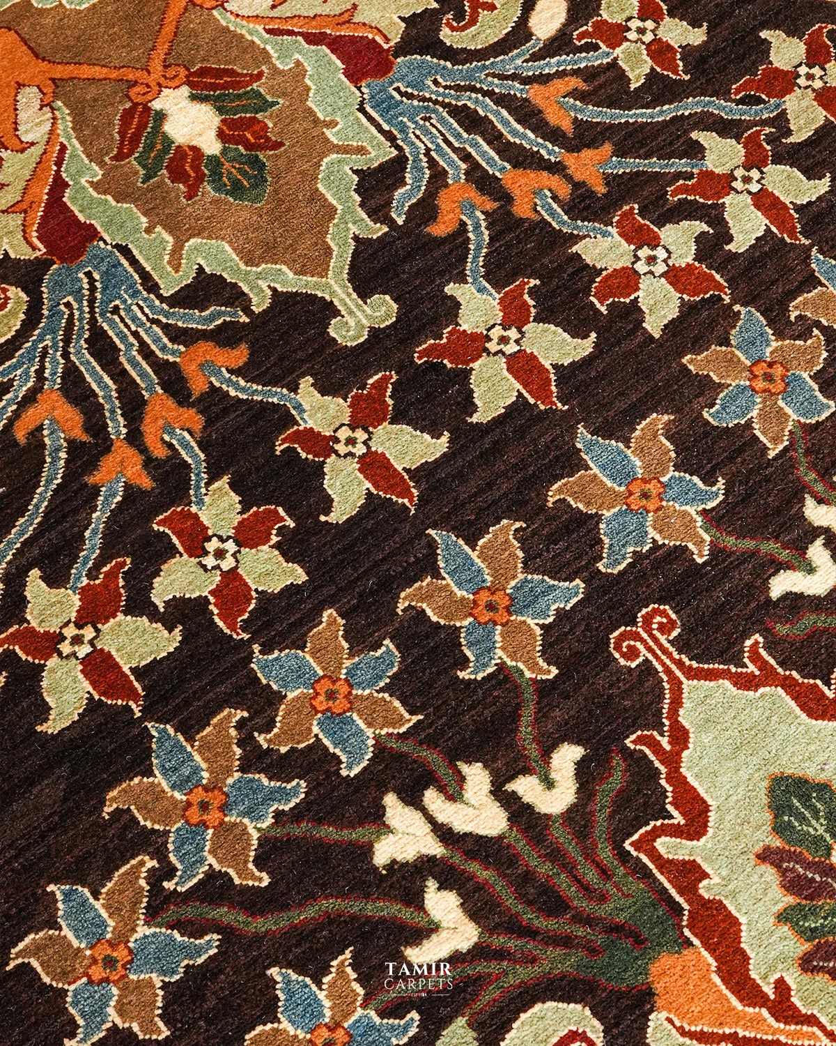 A close-up of a patterned area rug featuring a dark brown textured background with stylized floral motifs in rust red, light blue, beige, and green. A small 'TAMIR CARPETS' brand label is visible near the bottom center.