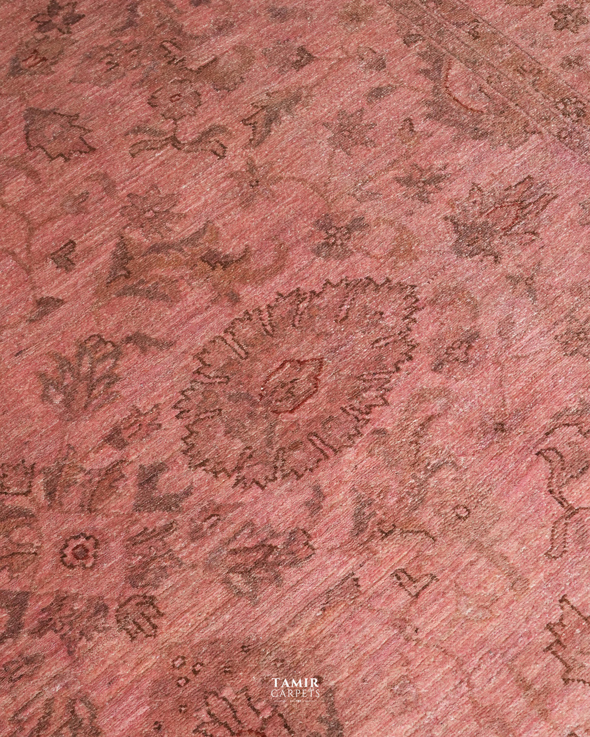 A close-up of a pinkish-red patterned rug with a traditional floral and medallion design in darker brown tones. The brand name 'TAMIR CARPETS' is visible as a watermark at the bottom center.