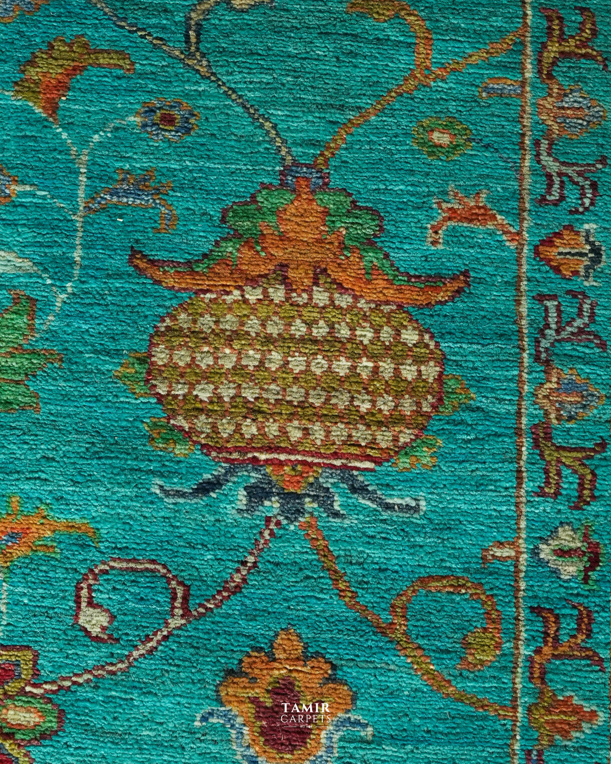 A close-up of a woven rug or textile with a turquoise background and an ornate central floral motif in brown, gold, green, and cream. The image is tightly cropped to show texture and pattern detail, with a small visible 'TAMIR CARPETS' watermark near the bottom center.