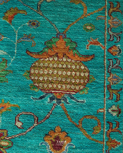 A close-up of a woven rug or textile with a turquoise background and an ornate central floral motif in brown, gold, green, and cream. The image is tightly cropped to show texture and pattern detail, with a small visible 'TAMIR CARPETS' watermark near the bottom center.
