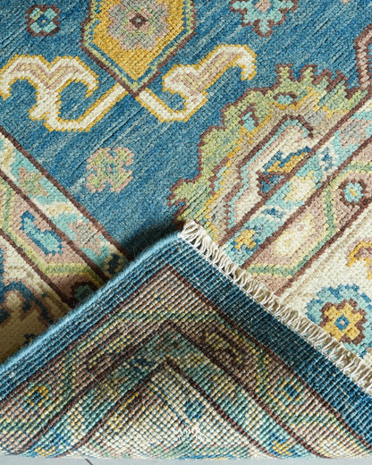 Close-up of a patterned area rug corner showing a blue field with cream, beige, yellow, and brown ornamental motifs, plus a fringed edge and the reverse side texture.