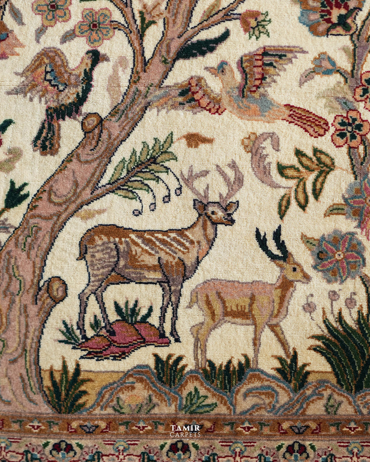 Close-up of a woven textile or rug pattern featuring deer, birds, floral motifs, and tree branches on an off-white background, with the brand name TAMIR CARPETS visible near the bottom.