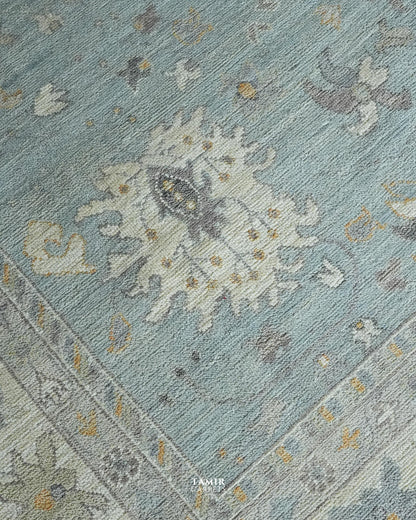 A close-up of a traditional area rug featuring a dusty blue field with a large central cream and gray floral medallion, accented with small gold and gray details, and a matching patterned border.