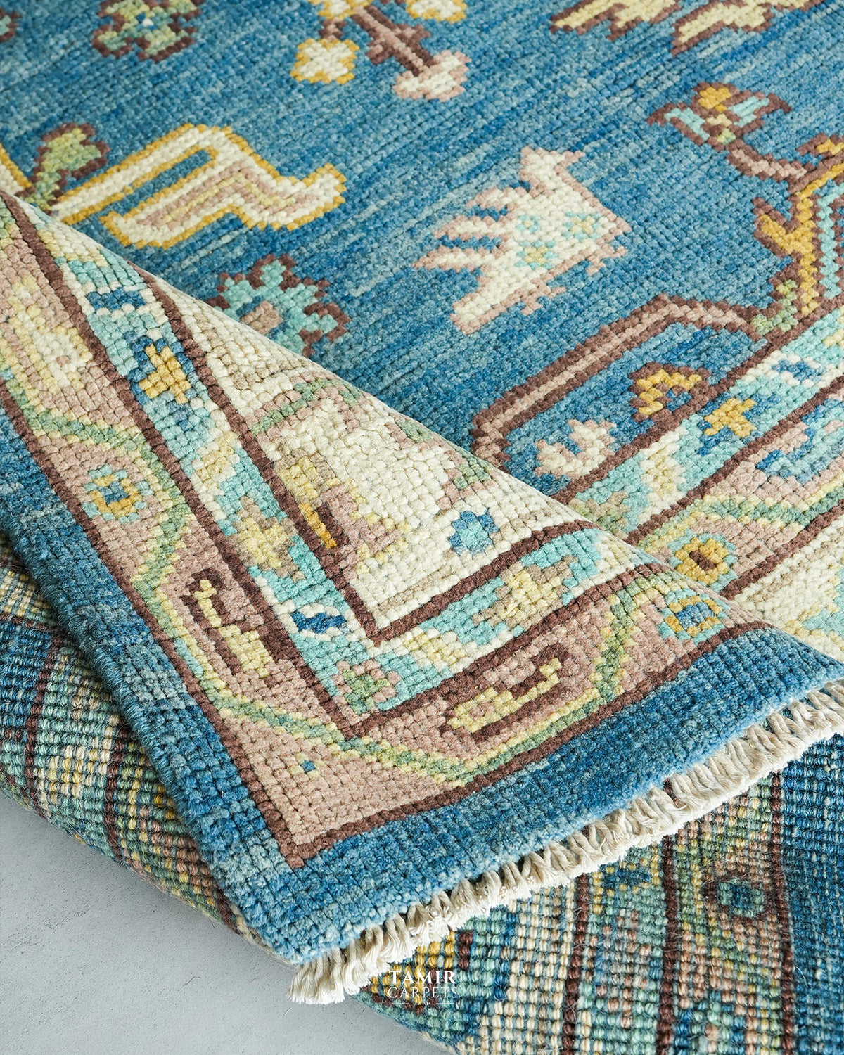 A close-up of a traditional handwoven rug with a folded corner showing its cream and brown border, beige fringe, and a blue field with abstract motifs.