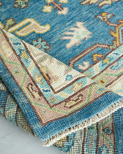 A close-up of a traditional handwoven rug with a folded corner showing its cream and brown border, beige fringe, and a blue field with abstract motifs.
