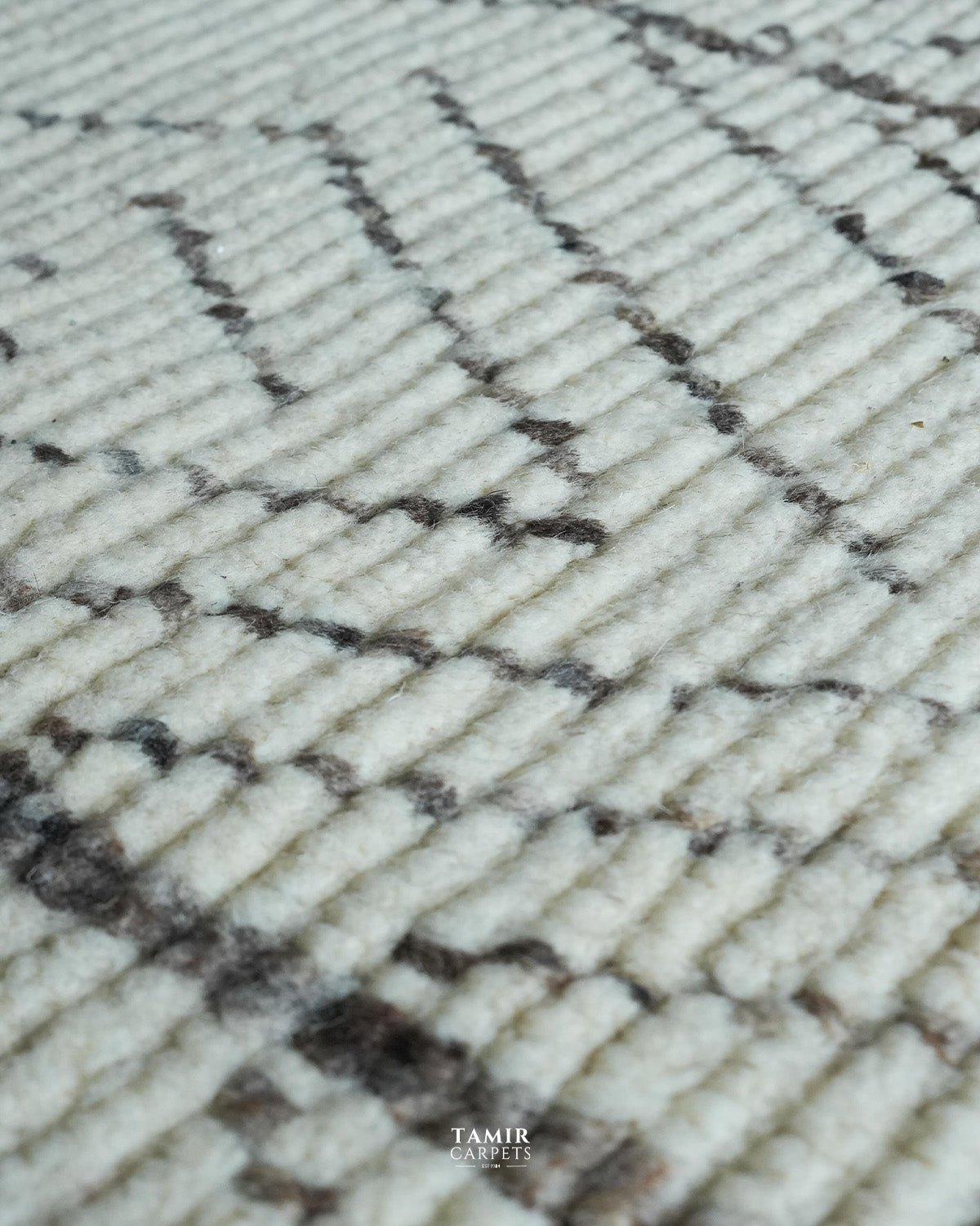 Close-up of a textured rug with a cream-colored base and a dark brown geometric zigzag pattern, featuring a 'TAMIR CARPETS' watermark at the bottom.