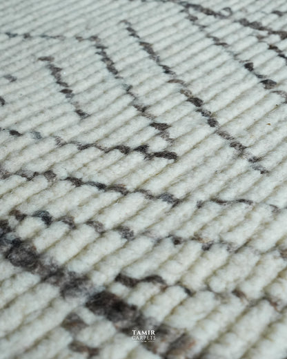Close-up of a textured rug with a cream-colored base and a dark brown geometric zigzag pattern, featuring a 'TAMIR CARPETS' watermark at the bottom.
