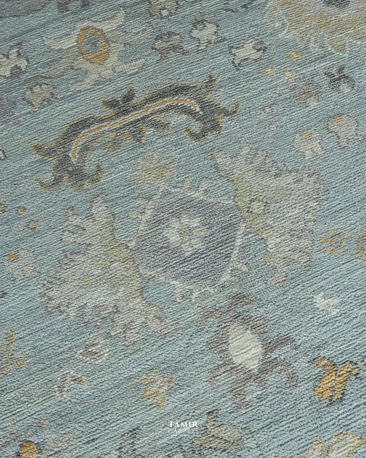 A close-up of a textured area rug featuring a faded, distressed traditional medallion pattern in muted shades of dusty blue, gray, beige, and gold.