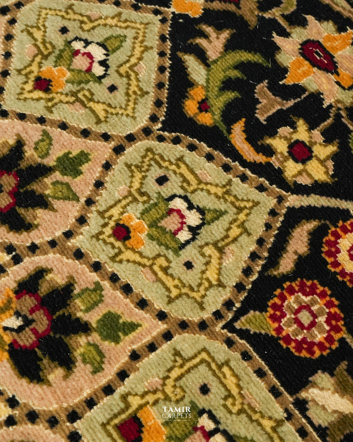 Close-up of a woven textile or rug pattern with repeating diamond-shaped medallions in beige, black, green, yellow, red, and white, plus a small TAMIR DESIGNS watermark near the bottom.
