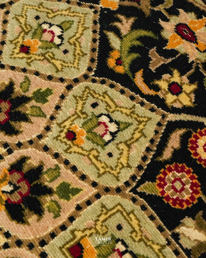 Close-up of a woven textile or rug pattern with repeating diamond-shaped medallions in beige, black, green, yellow, red, and white, plus a small TAMIR DESIGNS watermark near the bottom.