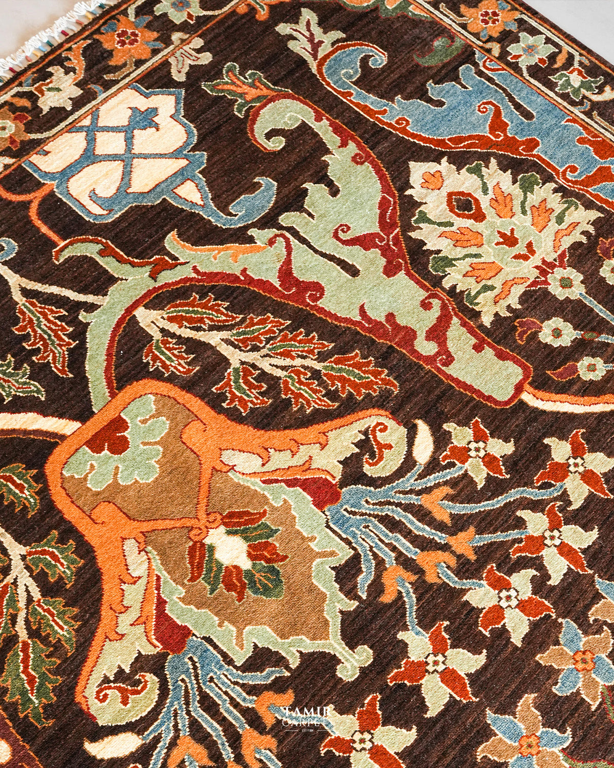 A close-up of a traditional patterned rug with a dark brown base and ornate floral and scroll motifs in beige, orange, light blue, and green.