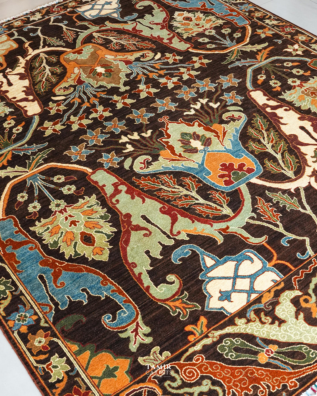 A close-up of an ornate area rug with a dark brown background and intricate floral and scroll motifs in beige, light green, orange, and blue.