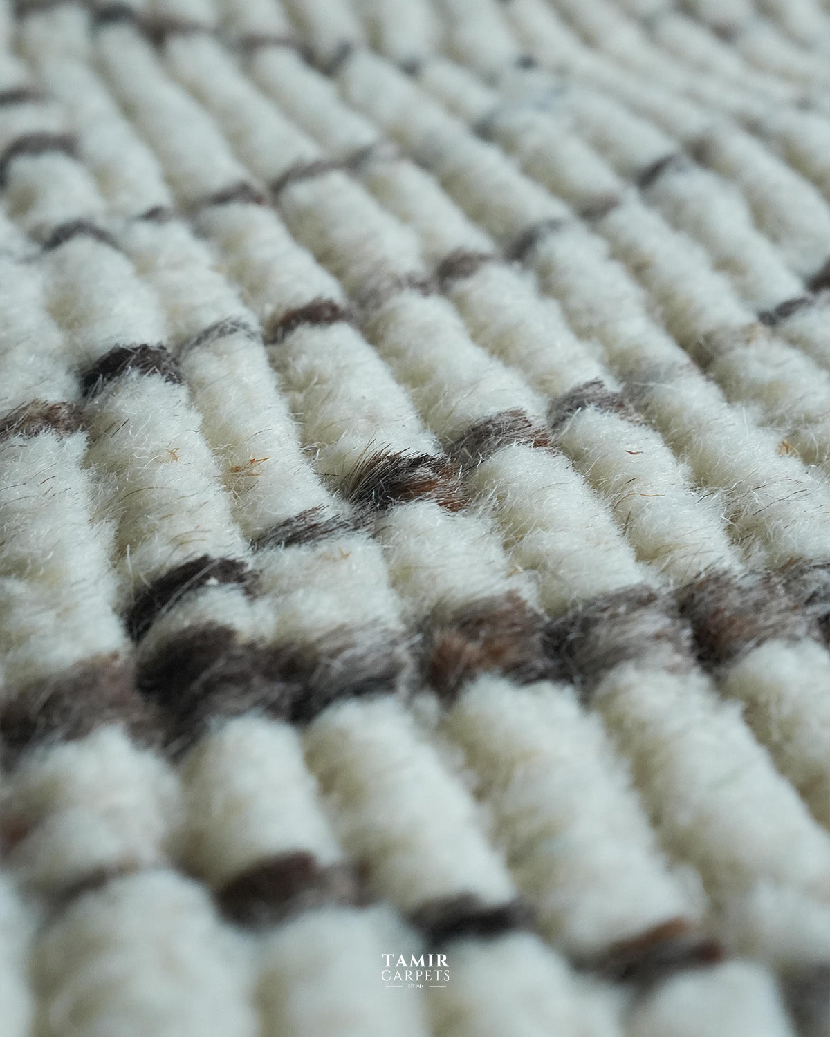 Close-up of a textured woven carpet with off-white loops and dark brown horizontal stripes, showing the pile and weave detail. A small 'TAMIR CARPETS' logo is visible at the bottom.