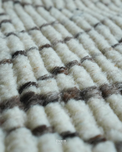 Close-up of a textured woven carpet with off-white loops and dark brown horizontal stripes, showing the pile and weave detail. A small 'TAMIR CARPETS' logo is visible at the bottom.